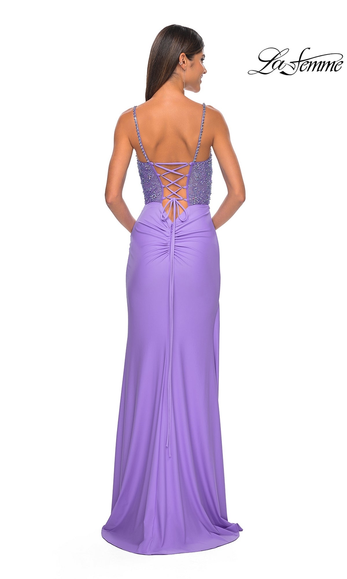 A woman stands facing away in the La Femme Beaded-Corset Long Prom Dress 32089, featuring a floor-length lavender design with thin straps, a beaded bodice, and lace-up back detailing.
